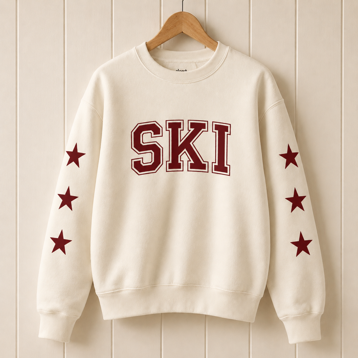 An off-white crewneck sweatshirt hanging on a wooden hanger against a cream-colored vertical wood paneled wall. The sweatshirt features the word 'SKI' in dark red varsity block letters outlined in white across the chest, and five dark red stars printed on each sleeve.