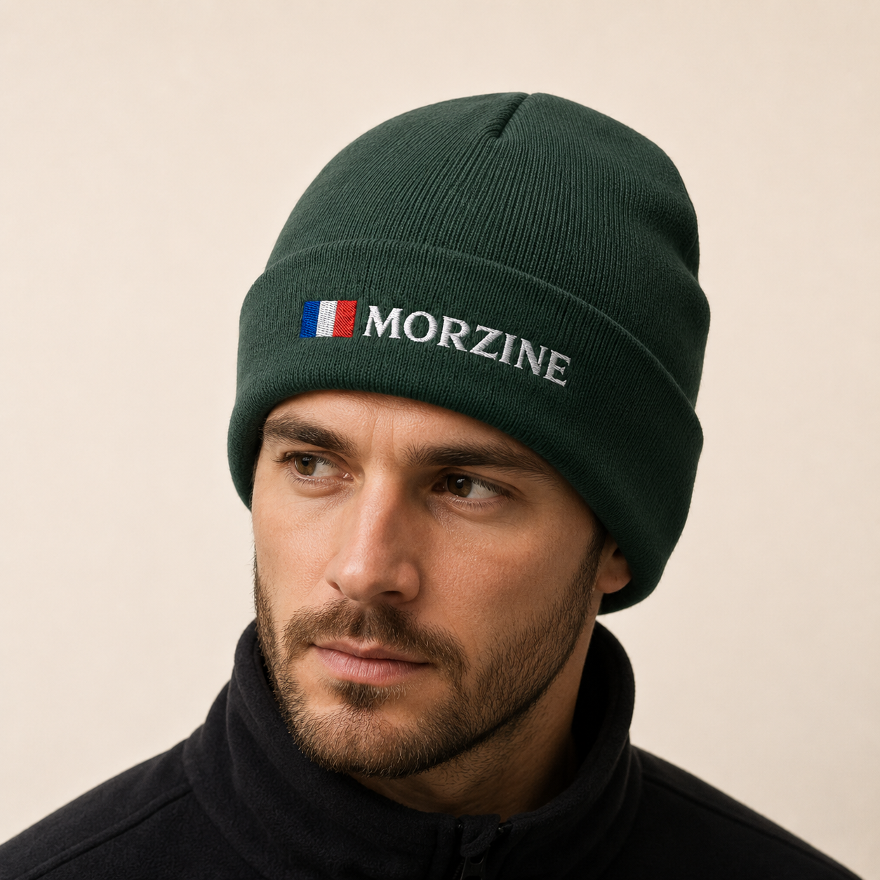 A man wearing a dark forest green knit beanie with 'MORZINE' embroidered in white on the front, next to a small French flag motif. The background is a solid, warm off-white studio backdrop.