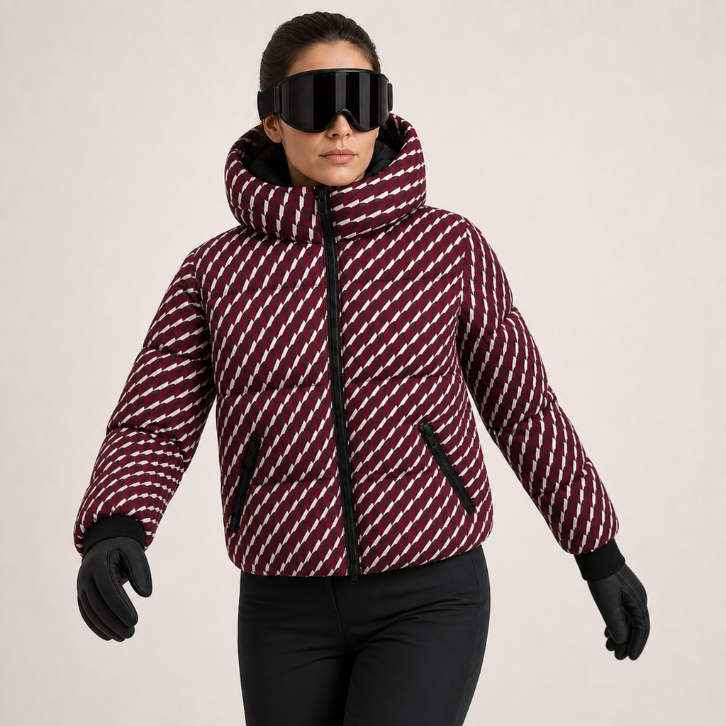 A woman wearing a patterned puffer jacket, black ski goggles, and black gloves, standing against a light beige studio background.
