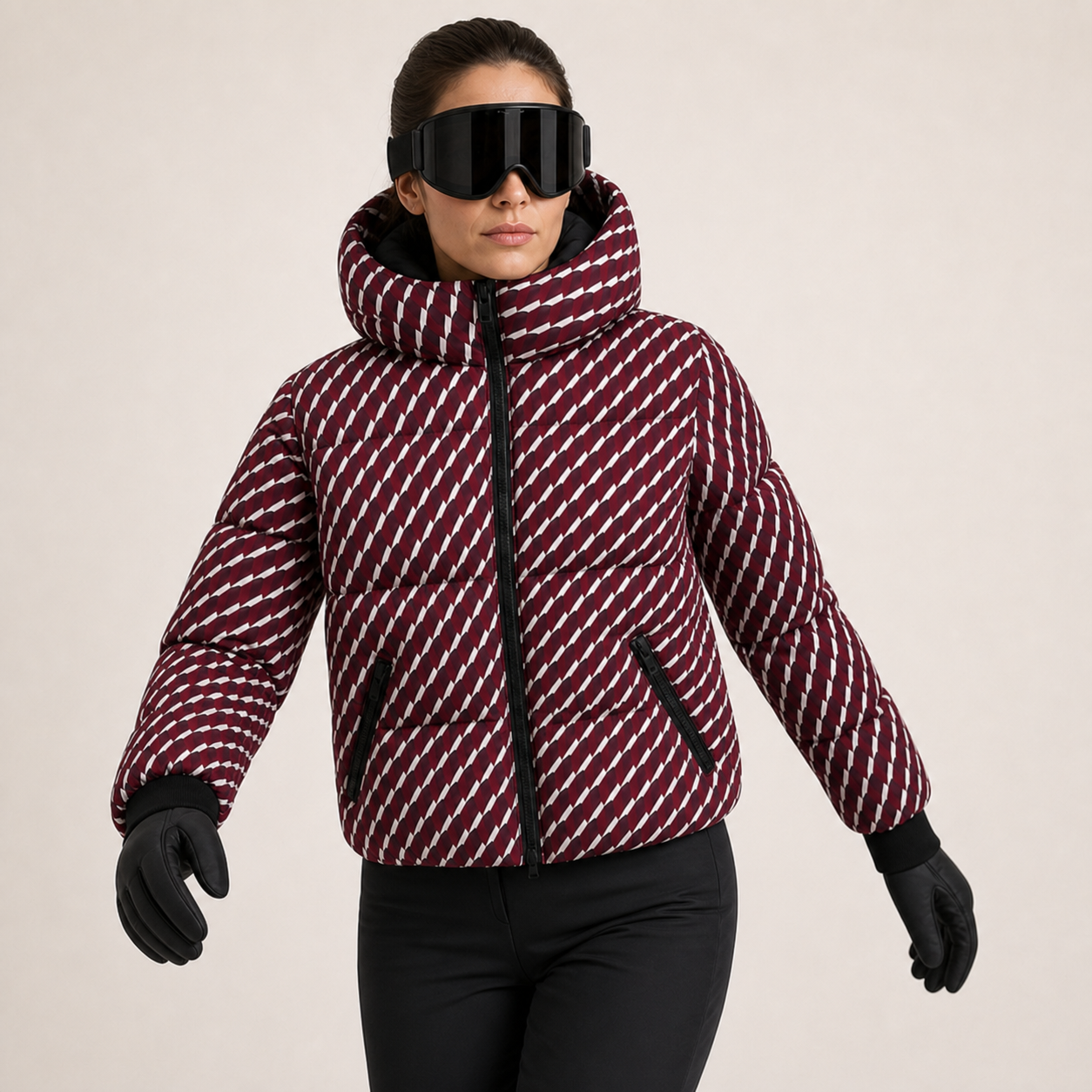 A woman wearing a patterned puffer jacket, black ski goggles, and black gloves, standing against a light beige studio background.