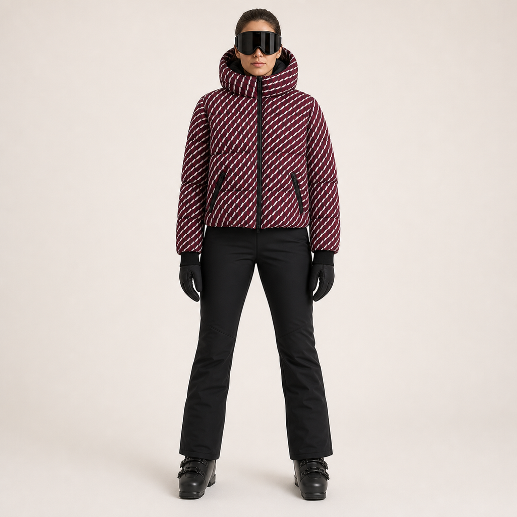 A model stands against a light beige studio background wearing a cropped puffer jacket with a diagonal striped pattern in burgundy and white, black ski pants, black gloves, and black ski goggles.
