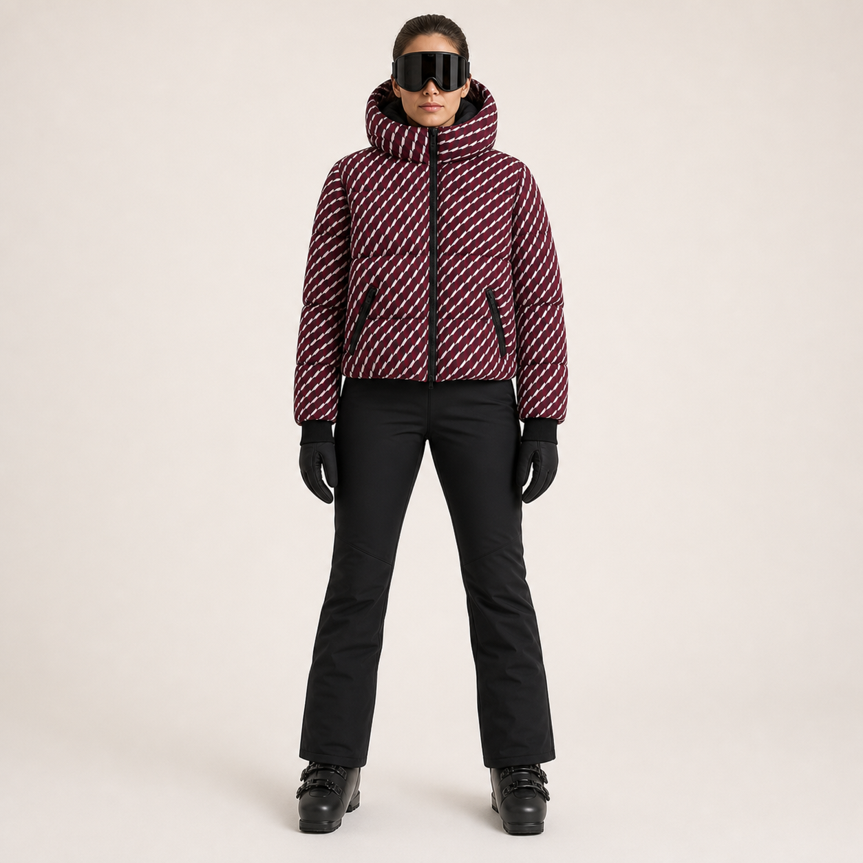 A model stands against a light beige studio background wearing a cropped puffer jacket with a diagonal striped pattern in burgundy and white, black ski pants, black gloves, and black ski goggles.