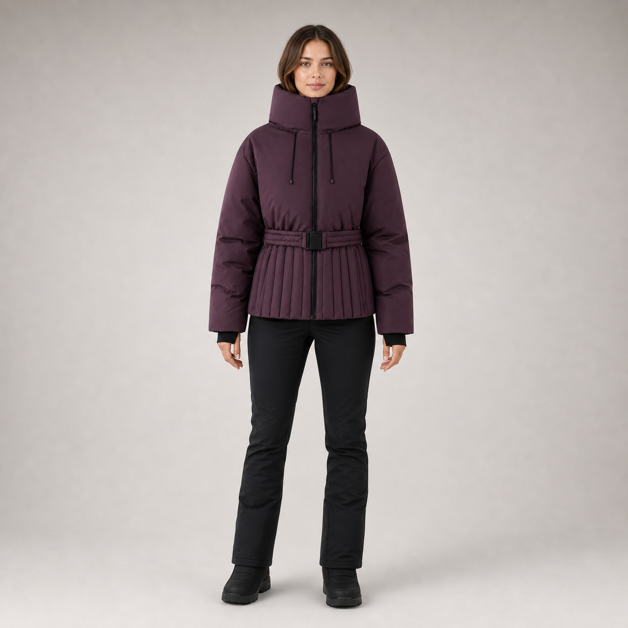A model stands against a light gray studio background wearing a dark plum purple puffer jacket with a high collar, drawstrings, a pleated front, and a matching belt, paired with black pants and boots.