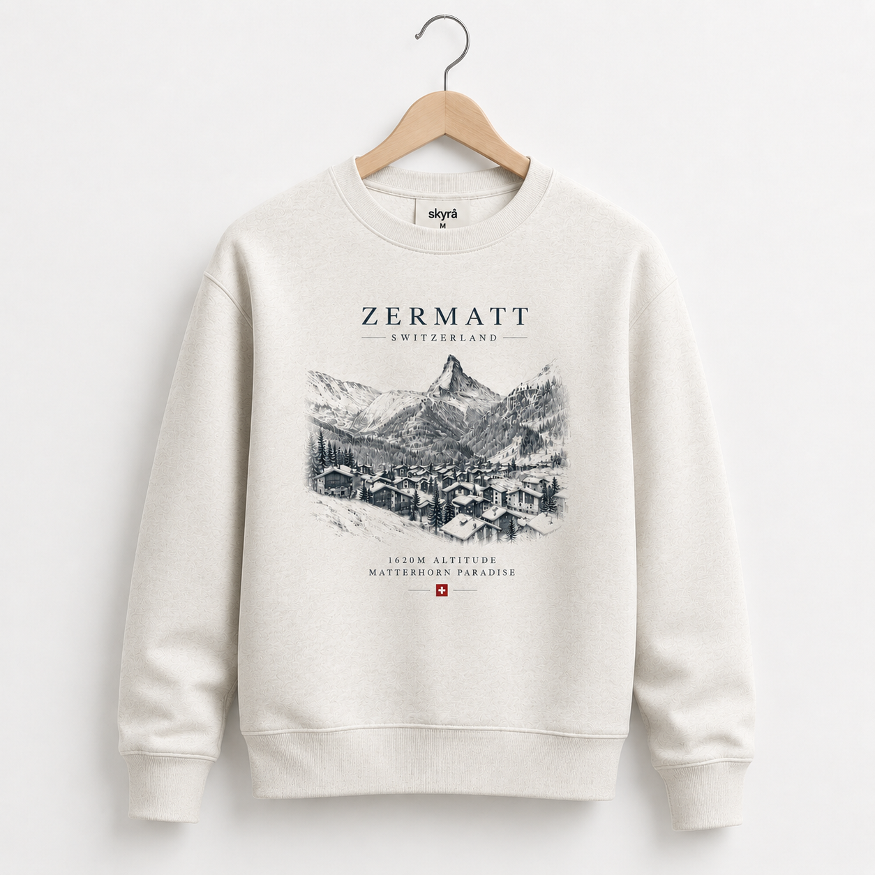 An off-white crewneck sweatshirt hanging on a wooden hanger against a light gray wall. The sweatshirt features a black line art graphic of Zermatt, Switzerland, with the text 'ZERMATT SWITZERLAND' and '1424M ALTITUDE MATTERING PARADISE' along with a small Swiss flag.