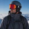 Video showing a rider standing on a snowy mountain wearing Skyra Performance Ski Goggles with a Red Mirror magnetic interchangeable lens and black strap. The rider is dressed in a dark ski jacket and helmet, with panoramic alpine peaks and clouds in the background. The reflective red lens highlights the goggles' premium design and mountain-ready performance in bright conditions.