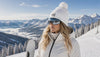 Female skier wearing a white Skyra pom pom beanie and ski goggles in a premium alpine ski resort surrounded by snow-covered mountains.